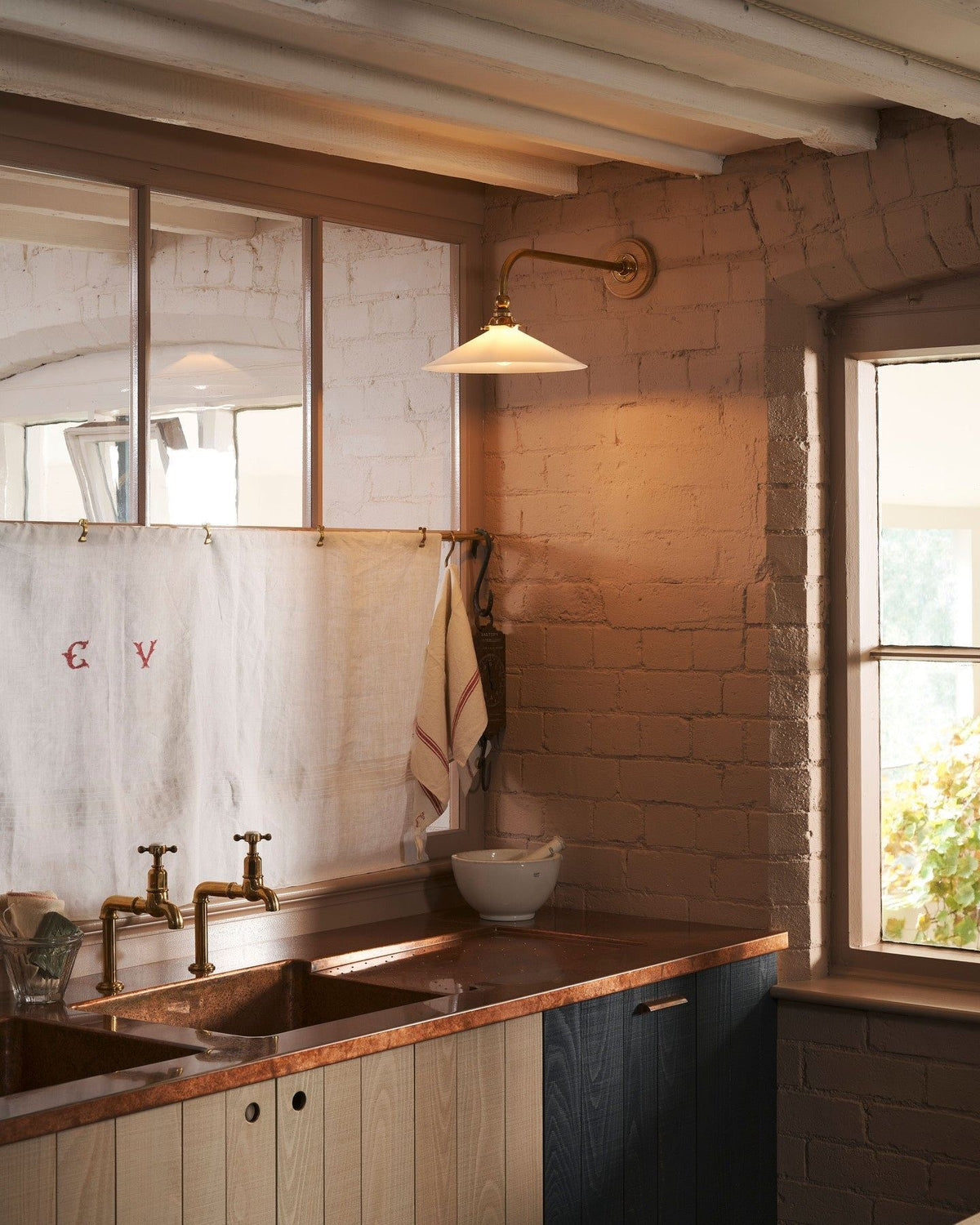 An oblique view of a large Victorian-inspired wall light with a brass arm and a milk glass shade fixed to a pink brick wall over modern rustic kitchen with an aged copper worktop. TEST