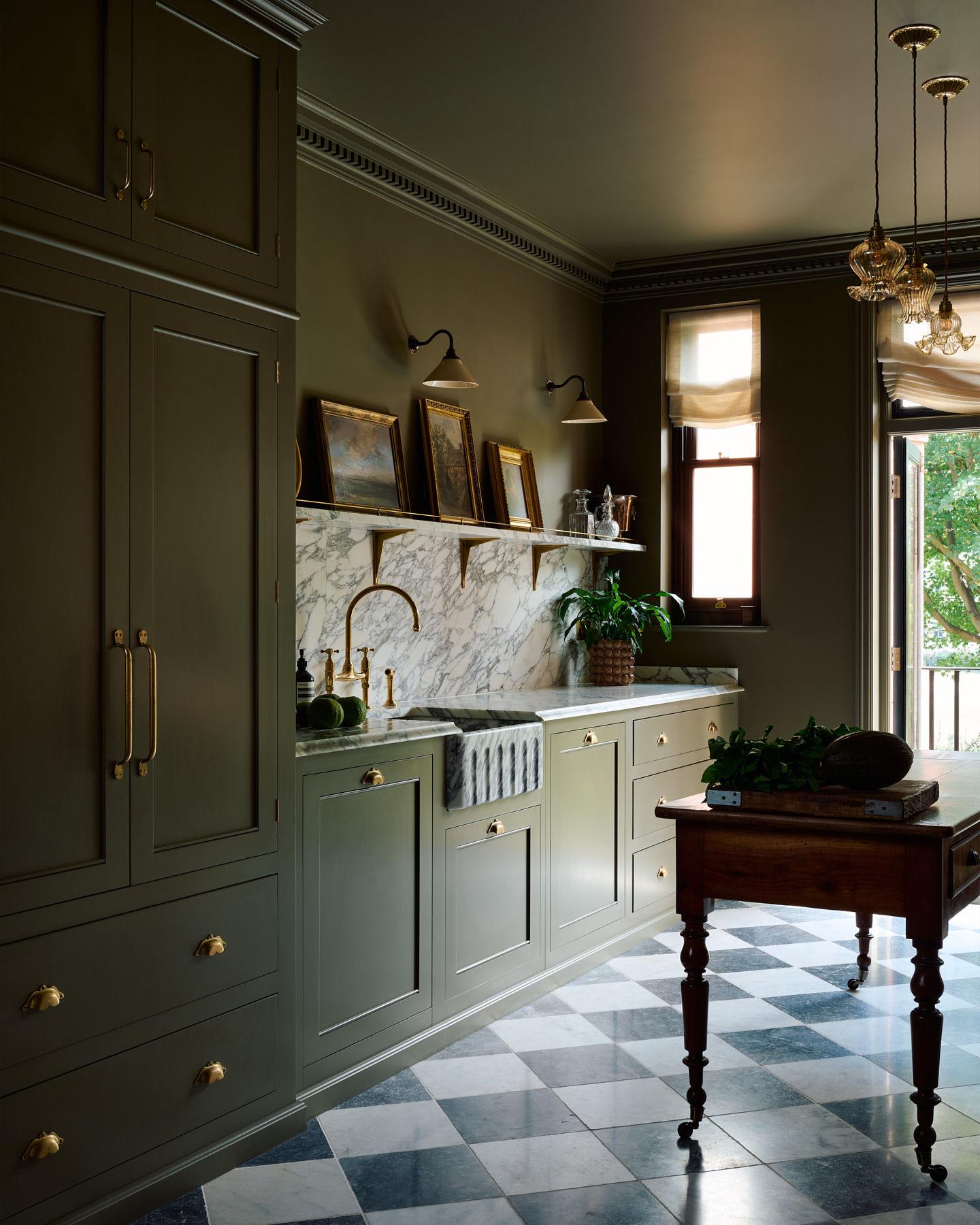 A straight-on view of a small wall light with an aged brass swan neck arm and a small cream glazed ceramic shade, fixed to a dark green wall above an Arabescato marble splashback and shelf, decorated with antique oil paintings in gilded frames.