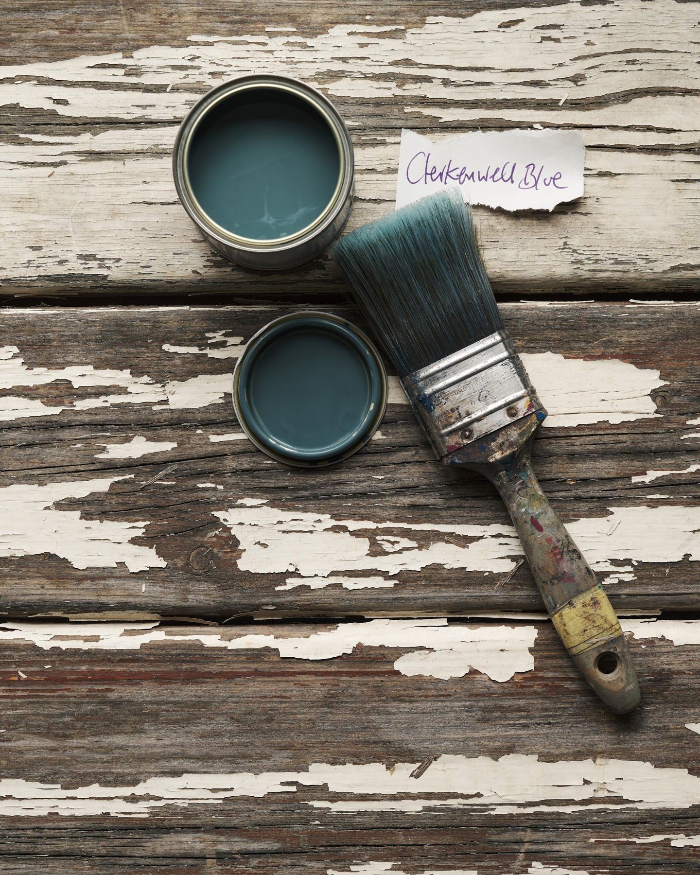 An overhead view of an open paint pot and a worn brush on a rustic tabletop. The green-tinged blue colour inside is labelled 'Clerkenwell Blue'.