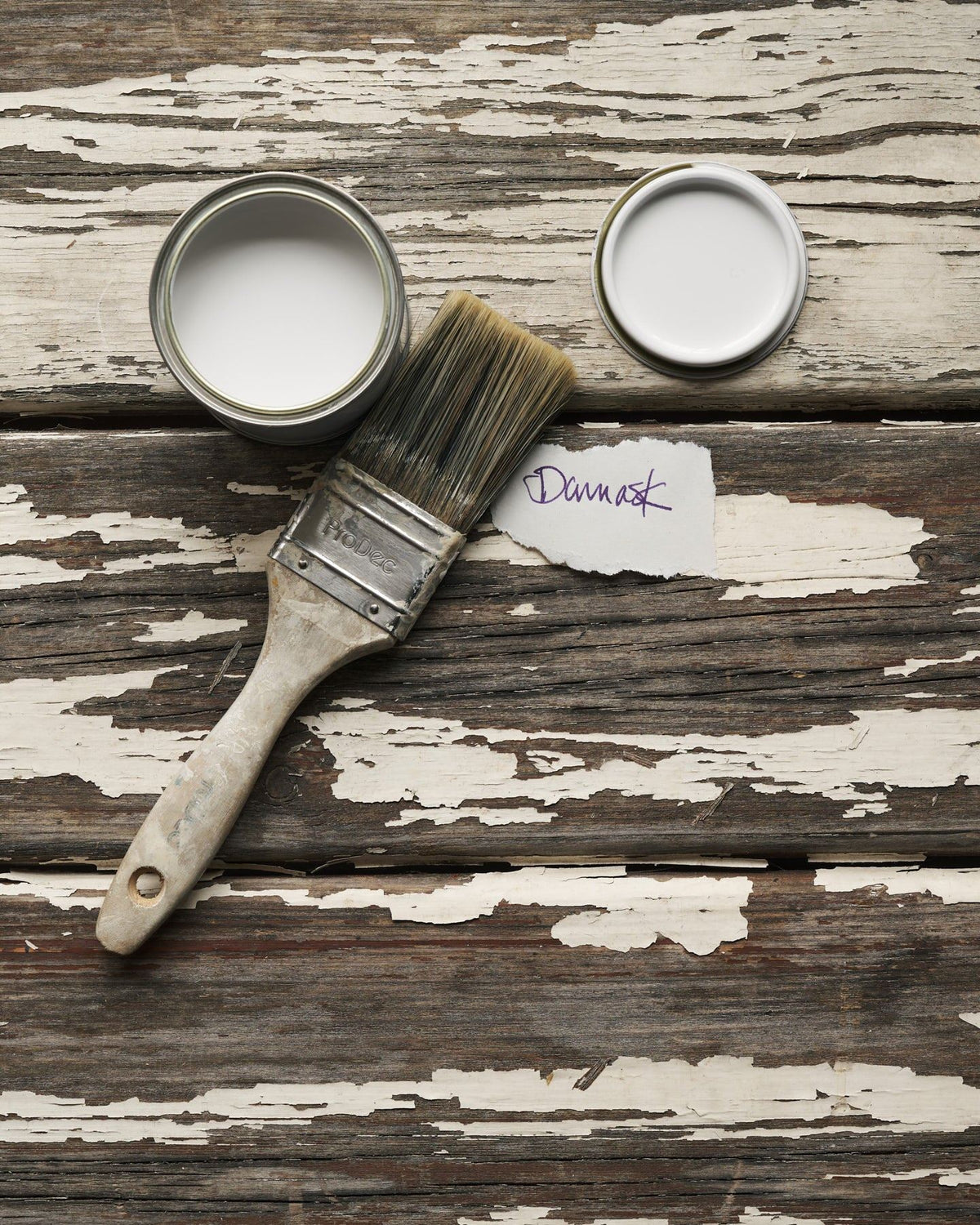 An overhead view of an open paint pot and a worn brush on a rustic tabletop. The off-white colour inside is labelled 'Damask'. TEST