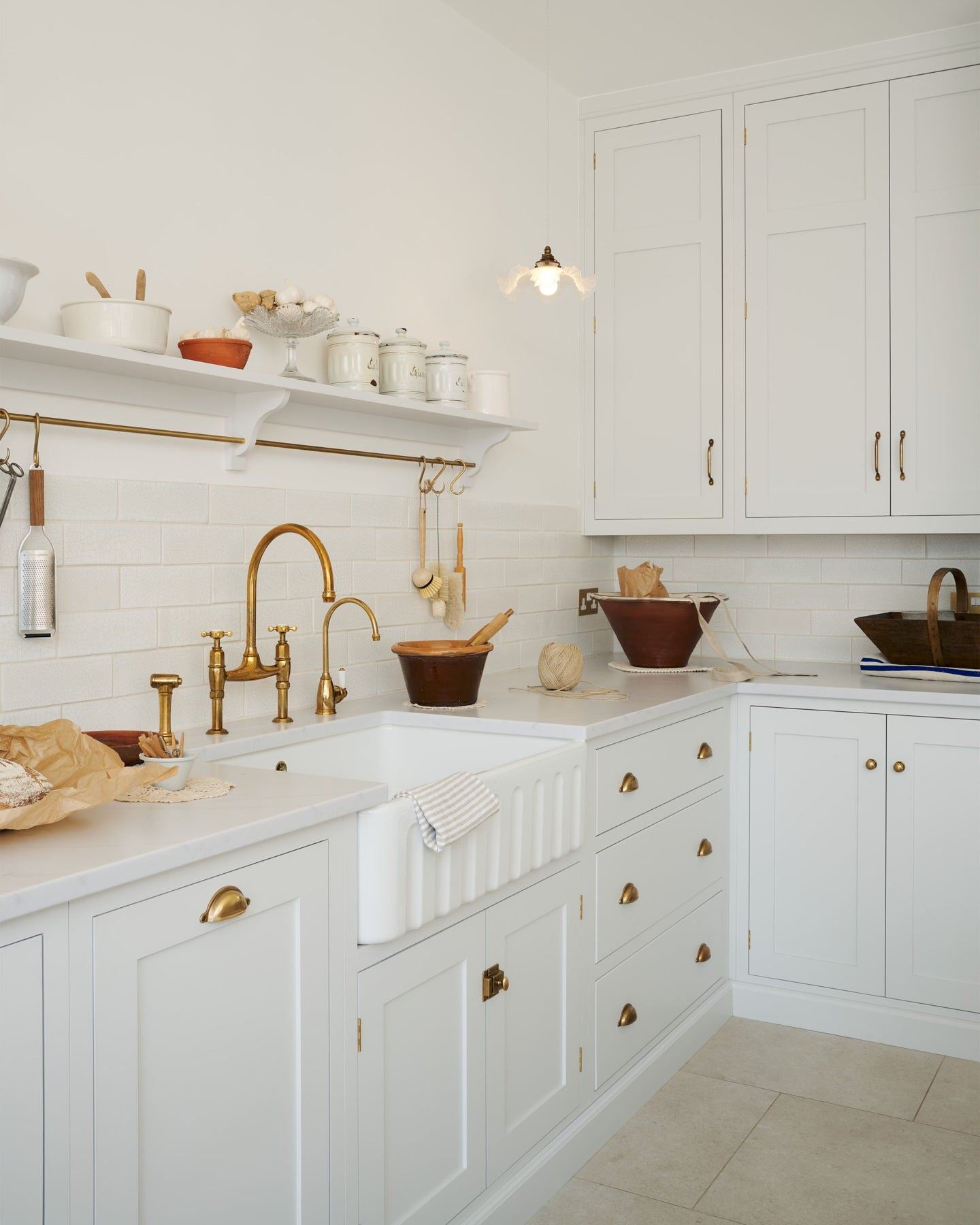 An oblique view of a simple Shaker-style kitchen with off-white painted cupboards, a fluted Belfast sink, aged brass taps and white metro tiles.