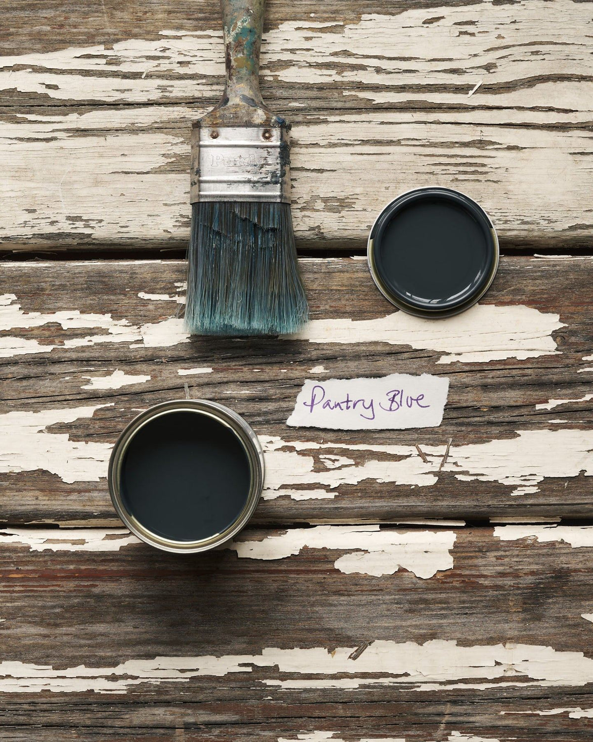 An overhead view of an open paint pot and a worn brush on a rustic tabletop. The dark blue colour inside is labelled 'Pantry Blue'. TEST
