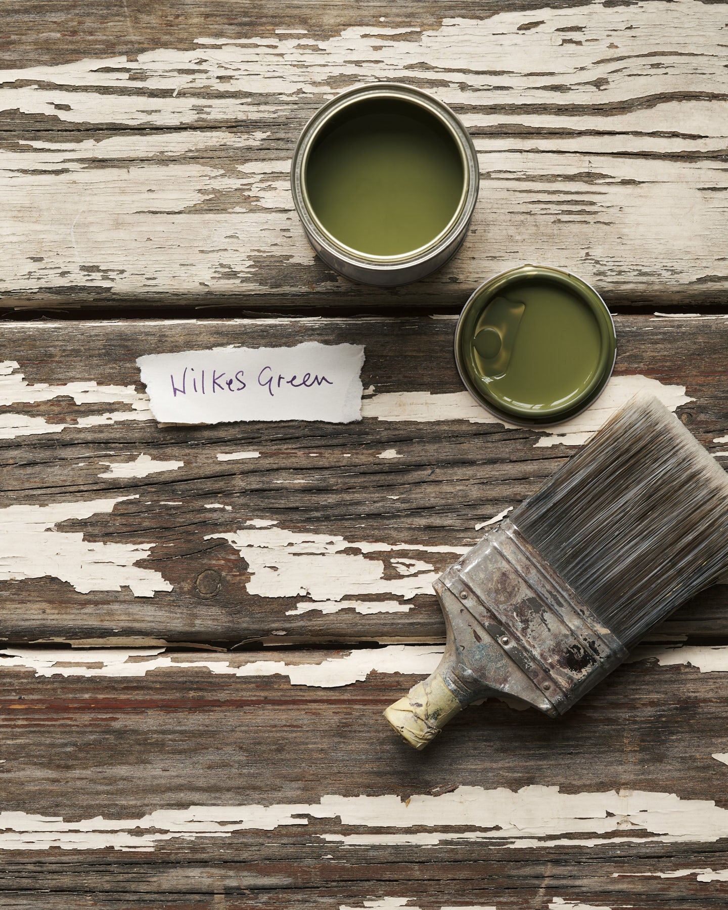 An overhead view of an open paint pot and a worn brush on a rustic tabletop. The mossy green colour inside is labelled 'Wilkes Green'.