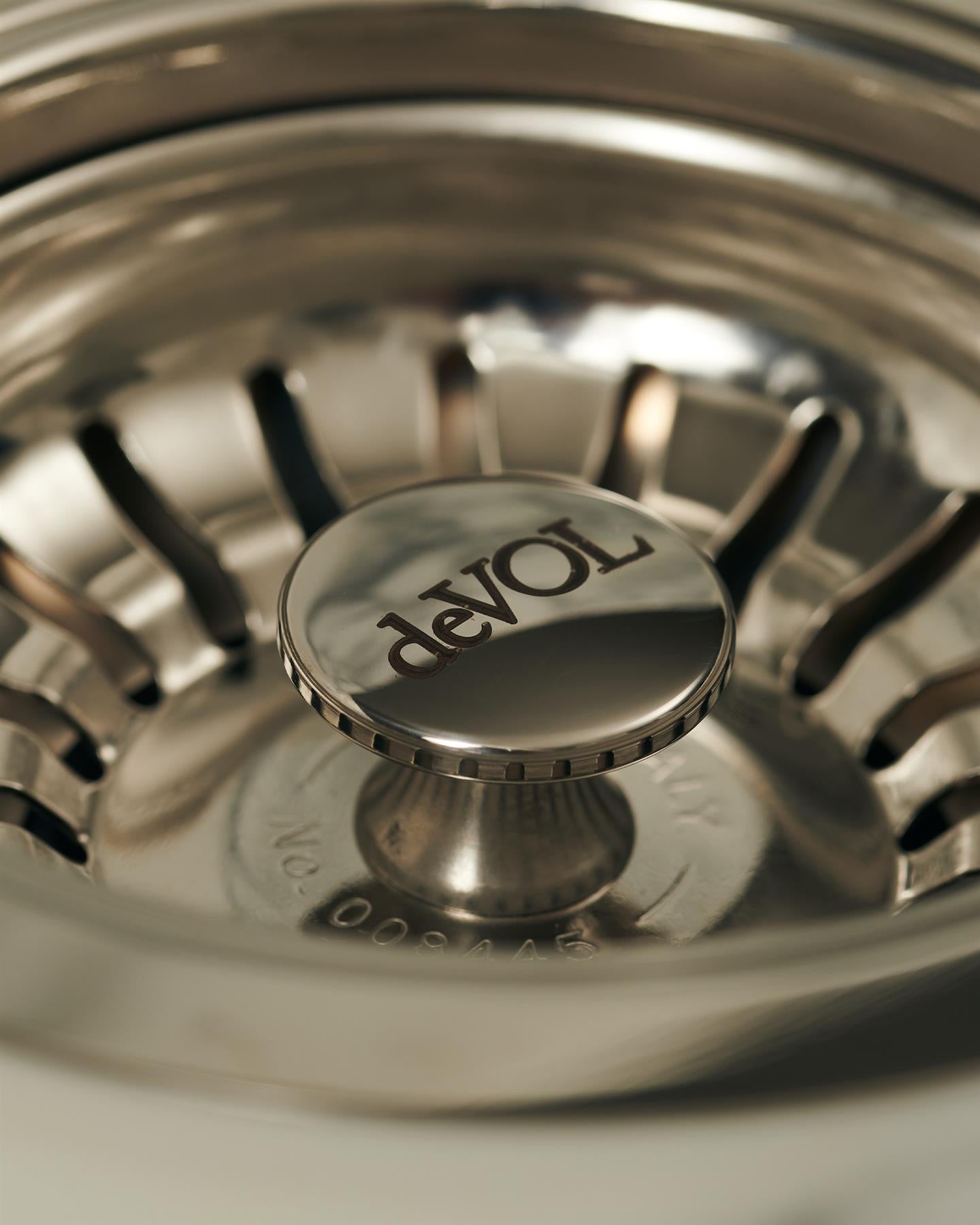 A close-up view of the central finial of a polished chrome basket waste strainer, there is a small 'deVOL' logo in the middle of the finial.