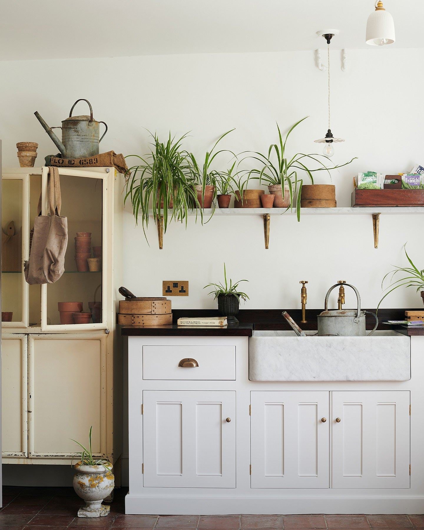 A straight-on view of a classic 13 amp twin plug socket in a mottled aged brass finish in a Mediterannean-inspired kitchen with a white painted Georgian-style sink cabinet, dark hardwood worktop, Carrara marble sink and a shelf full of lush spider plants.