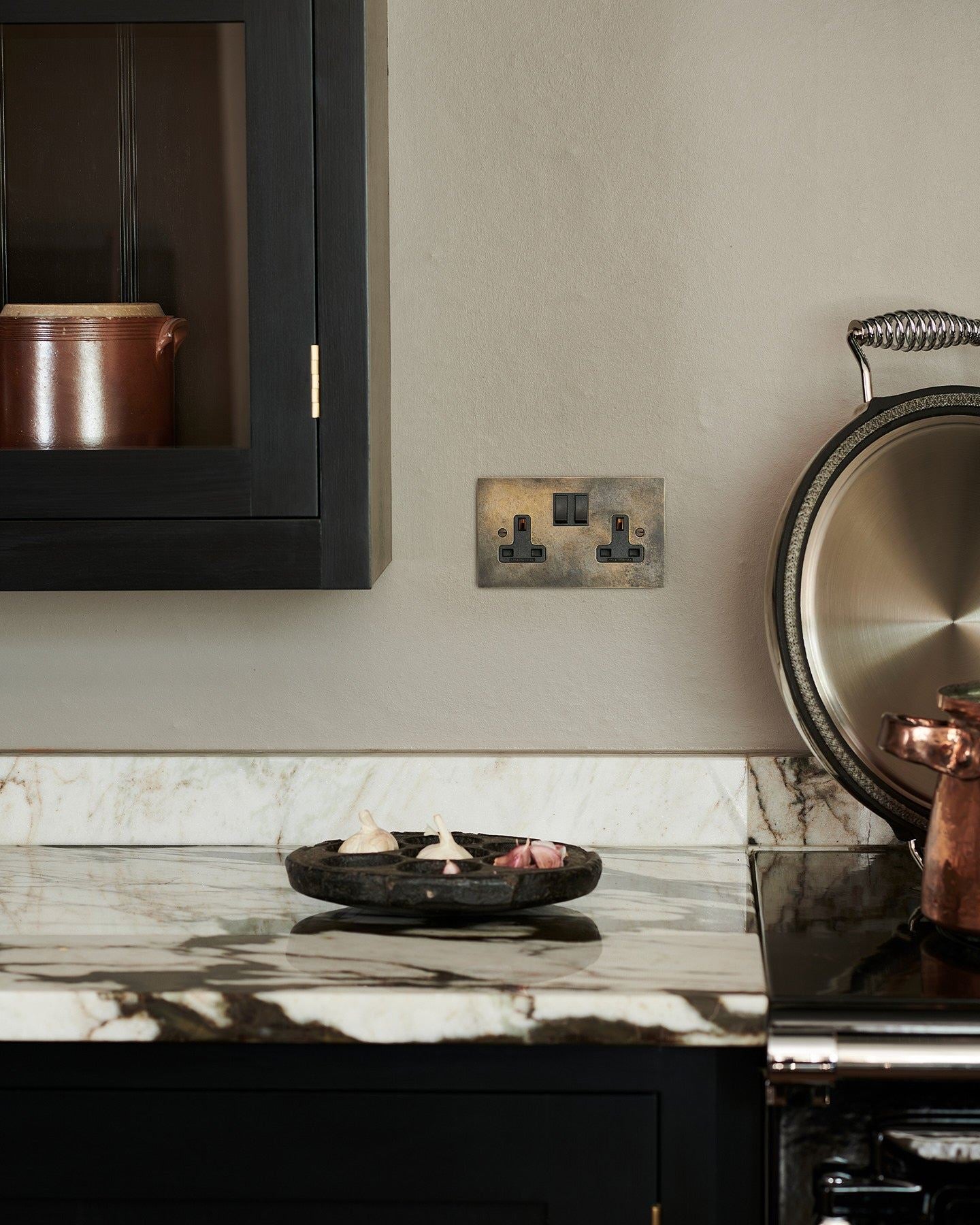 A straight-on view of a classic 13 amp twin plug sockets in a dark oxidised brass finish on a grey wall above a dramatic veined quartz worktop and black painted Shaker-style kitchen cabinets.
