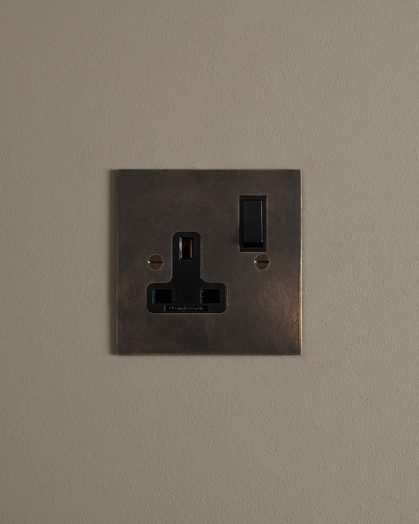 A straight-on view of a classic 13 amp single plug socket in a dark oxidised brass finish with black plastic switches.