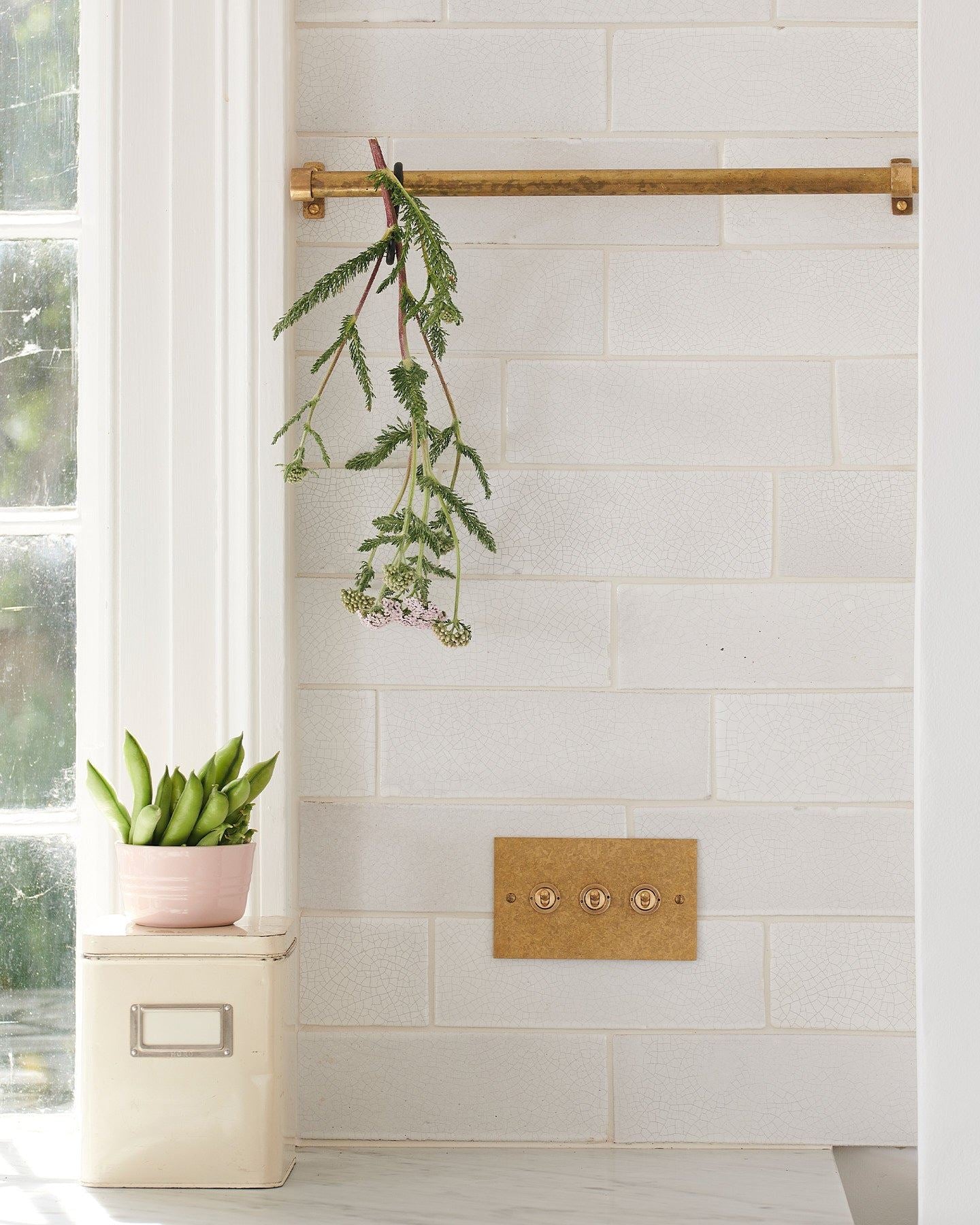 A straight-on view of a 3 gang classic toggle switch in an aged brass finish on white metro tiles with a small olive branch dangling from a short aged brass hanging rail above.