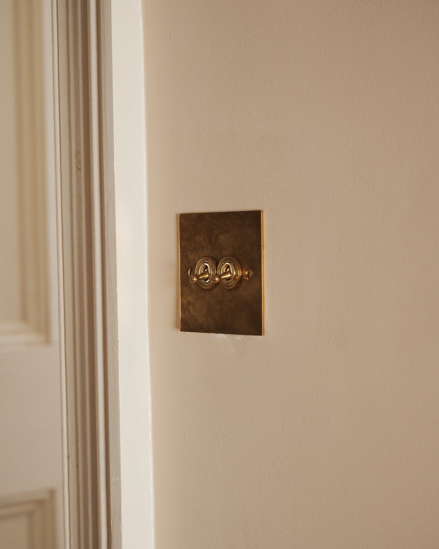 An oblique view of a 2 gang classic toggle switch in a warm burnished brass finish on soft pink walls with light painted woodwork.