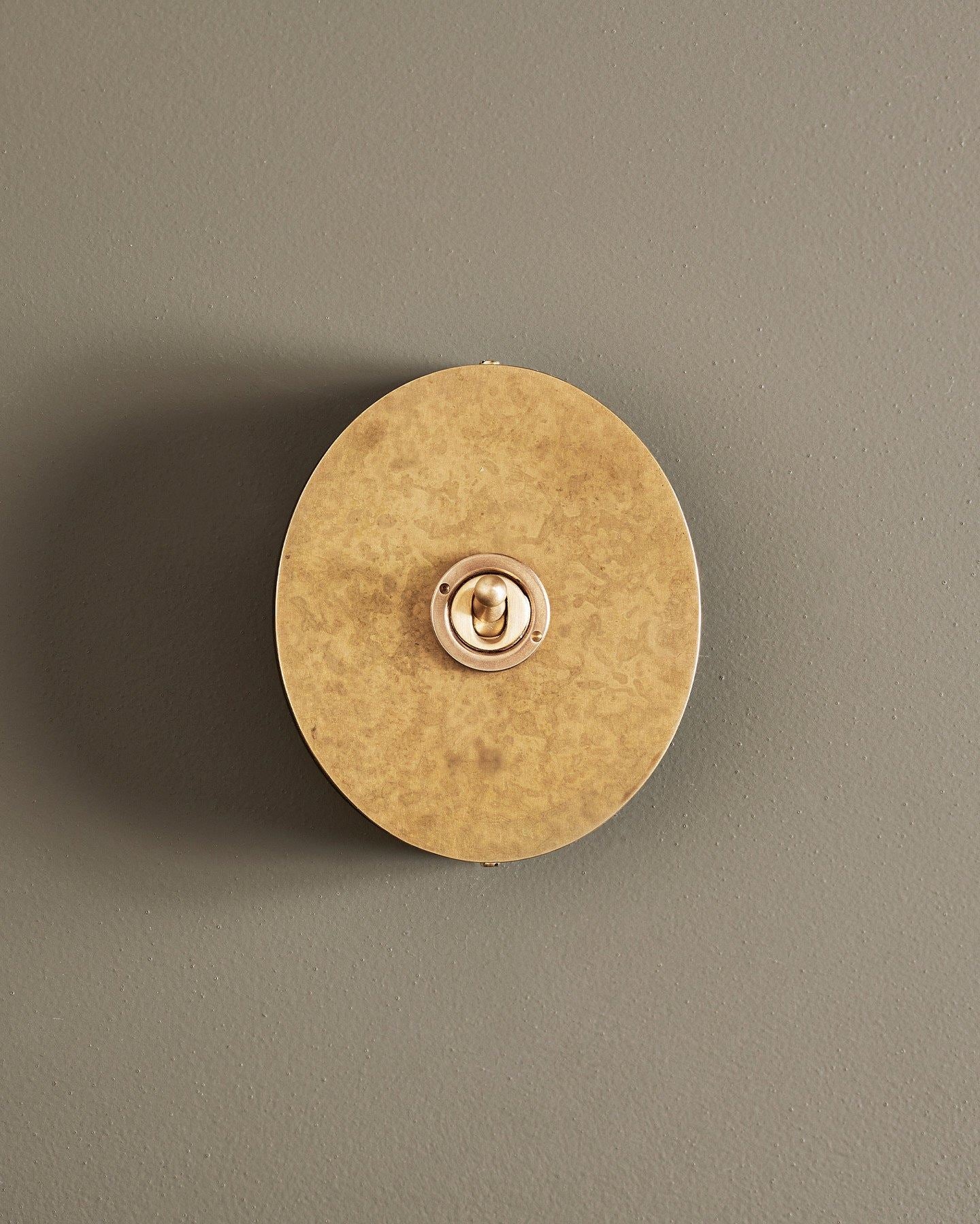 A straight-on view of a 1 gang oval-shaped toggle switch in a mottled aged brass finish with a black base.