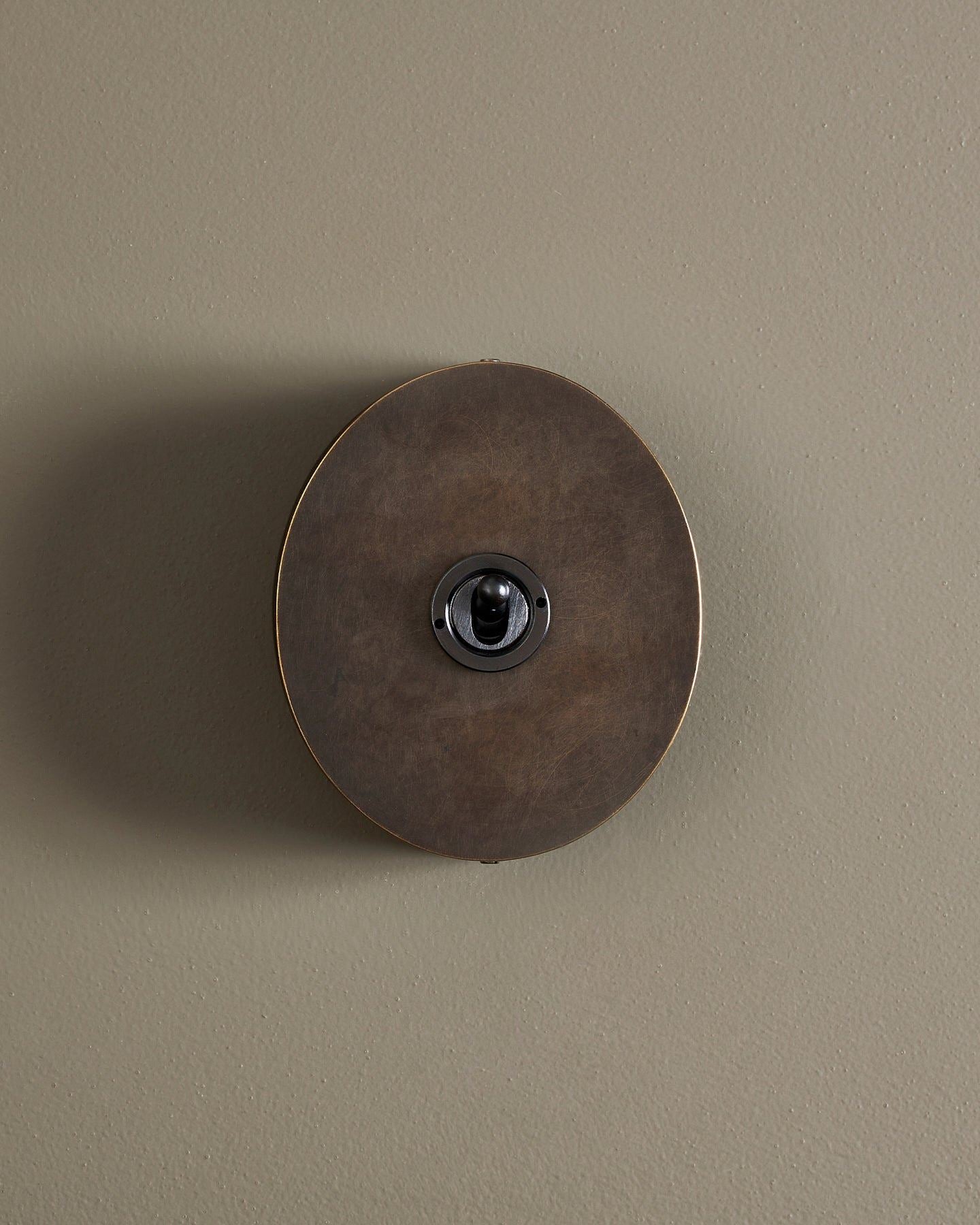 A straight-on view of a 1 gang oval-shaped toggle switch in a dark oxidised brass finish with a black base.