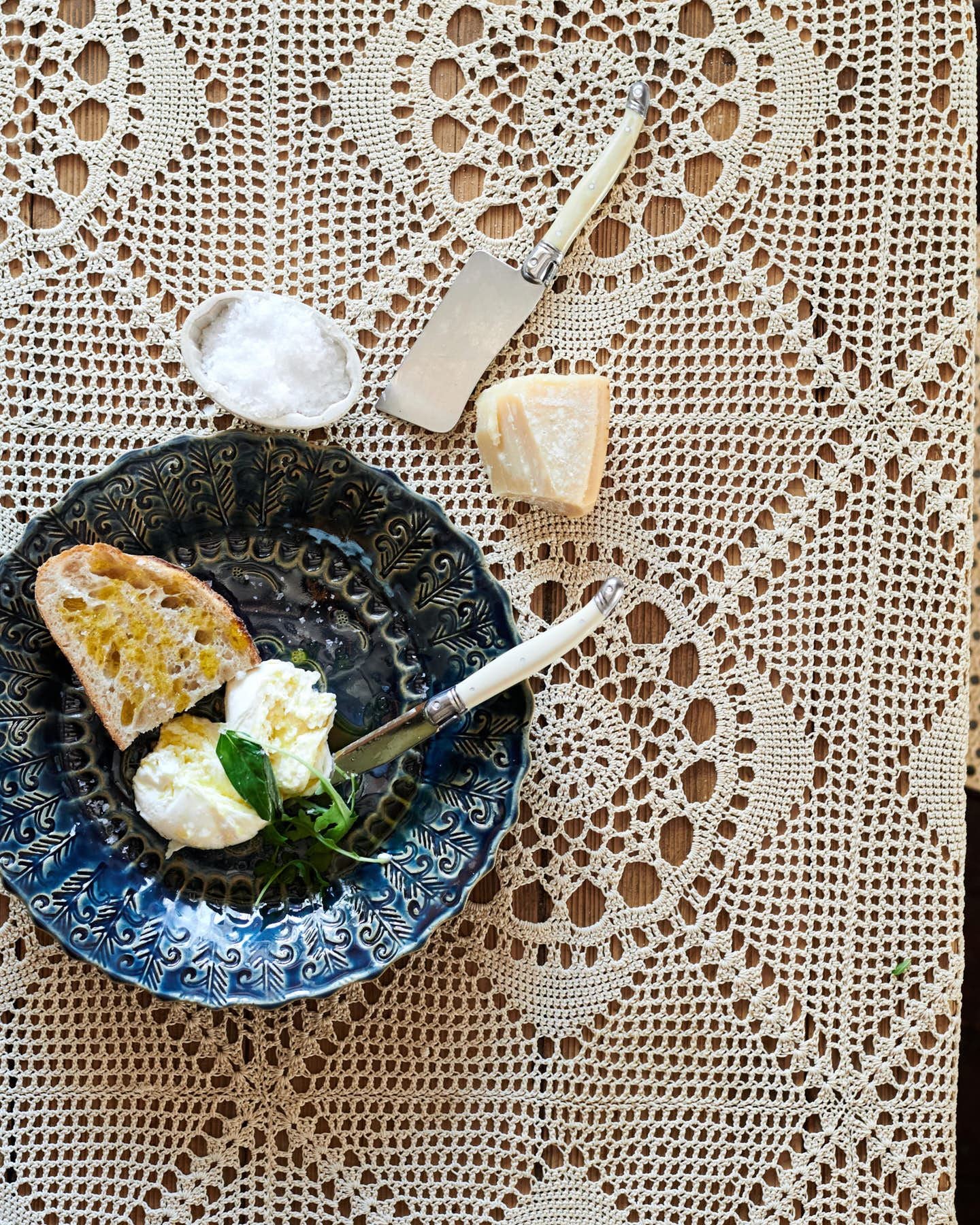 An overhead view of a round handmade bohemian-style blue ceramic platter with vintage silverware, cheese and homemade bread on a beautiful crochet lace tablecloth.