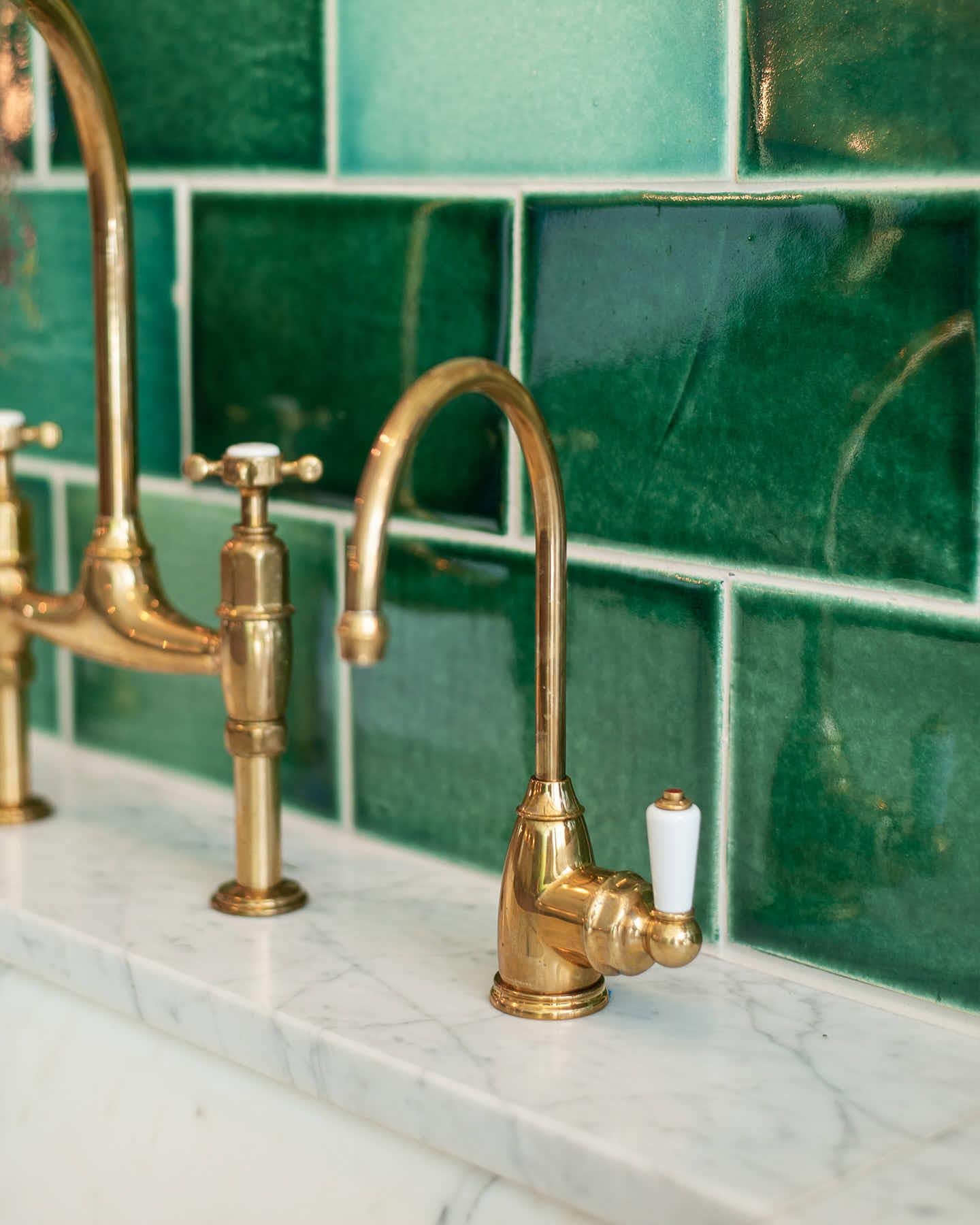 A small traditional-style aged brass tap with a white ceramic lever with glossy emerald green wall tiles behind