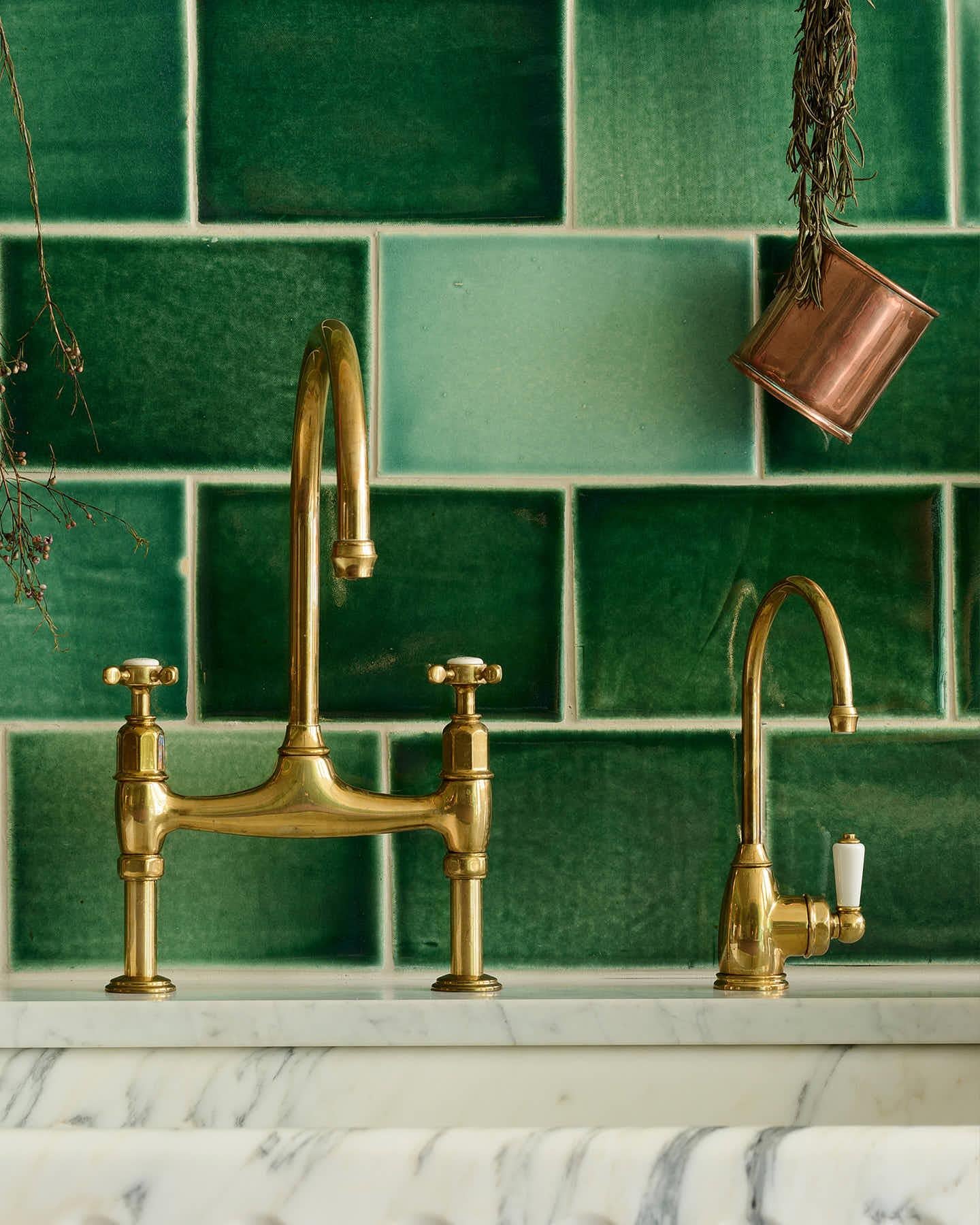 A straight-on view of a small traditional-style aged brass tap beside a larger aged brass mixer tap, with glossy emerald green wall tiles behind.
