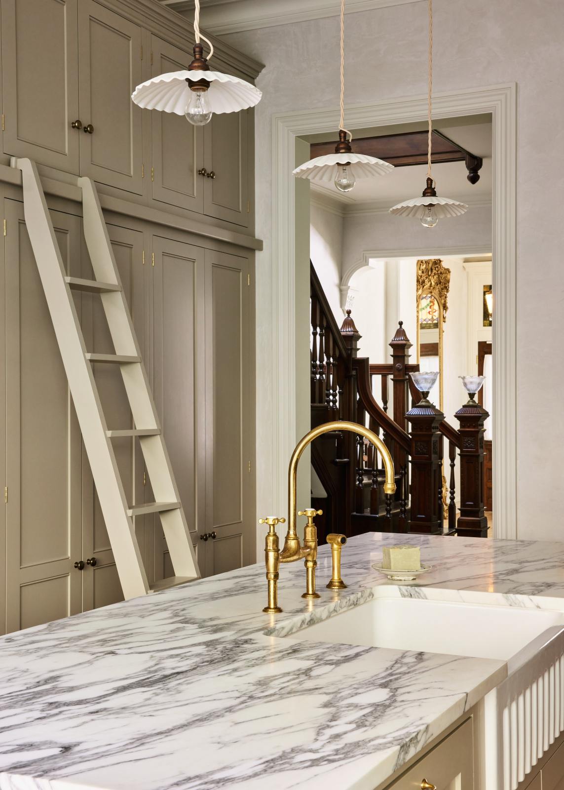An angled look at an elegant mushroom-brown, classic English kitchen features a large kitchen island with a fluted Belfast sink, an aged brass Ionian tap, Arabescato marble worktop, and a full-height pantry cupboard with a ladder. Beautifully decorated with artisan ceramic, narrow pleated porcelain lights, and a bar of hand-made soap, it adds a sophisticated touch to the elegant kitchen.