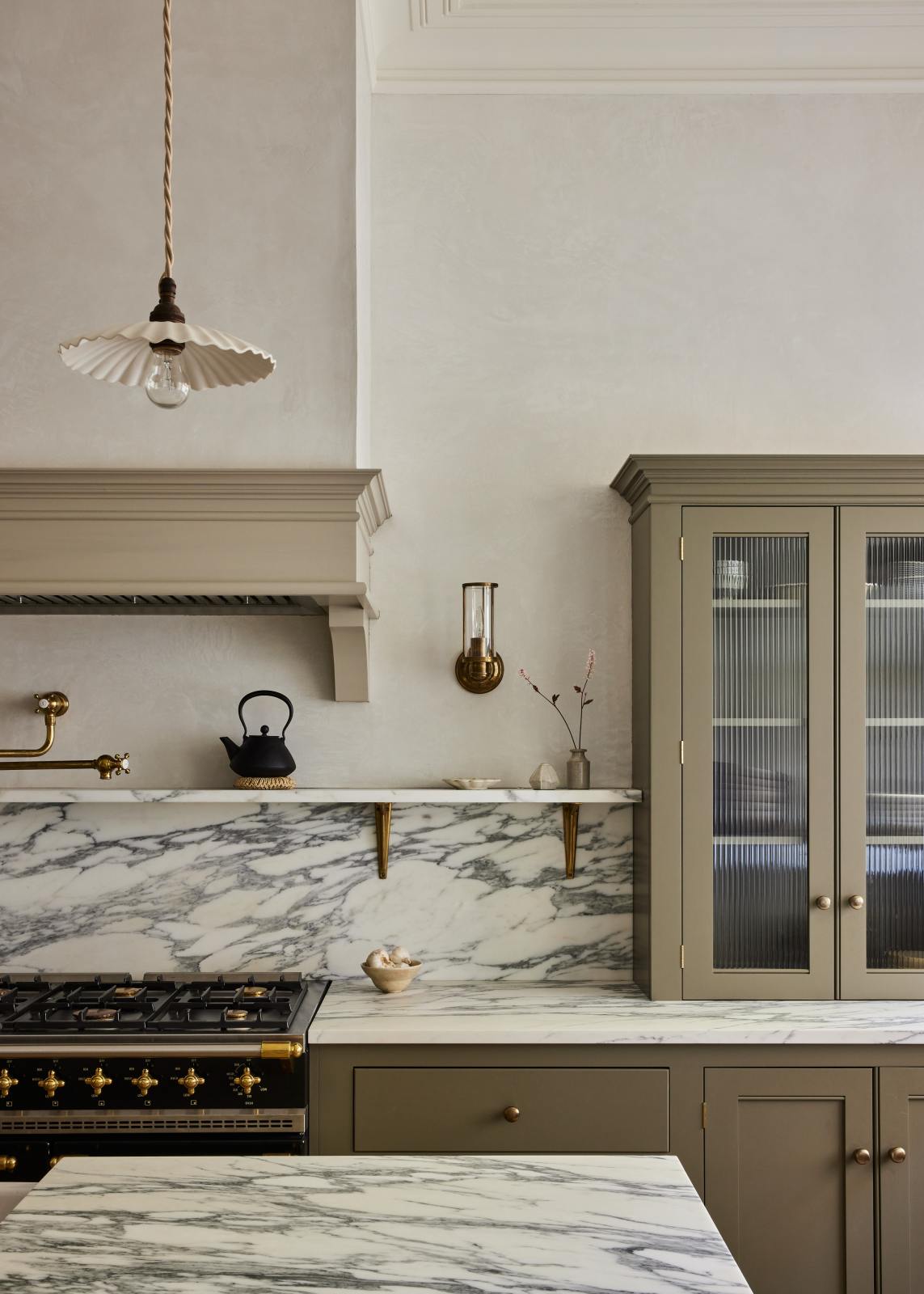 An elegant mushroom-brown, classic English kitchen features a cooker run with an Arabescato marble worktop, splashback, and shelf; a reeded countertop cupboard; a full-height pantry cupboard; and a large kitchen island with a fluted Belfast sink and an aged brass Ionian tap. Beautifully decorated with artisan ceramics, narrow pleated porcelain light, and a cast-iron teapot, it adds a sophisticated touch to the elegant kitchen.