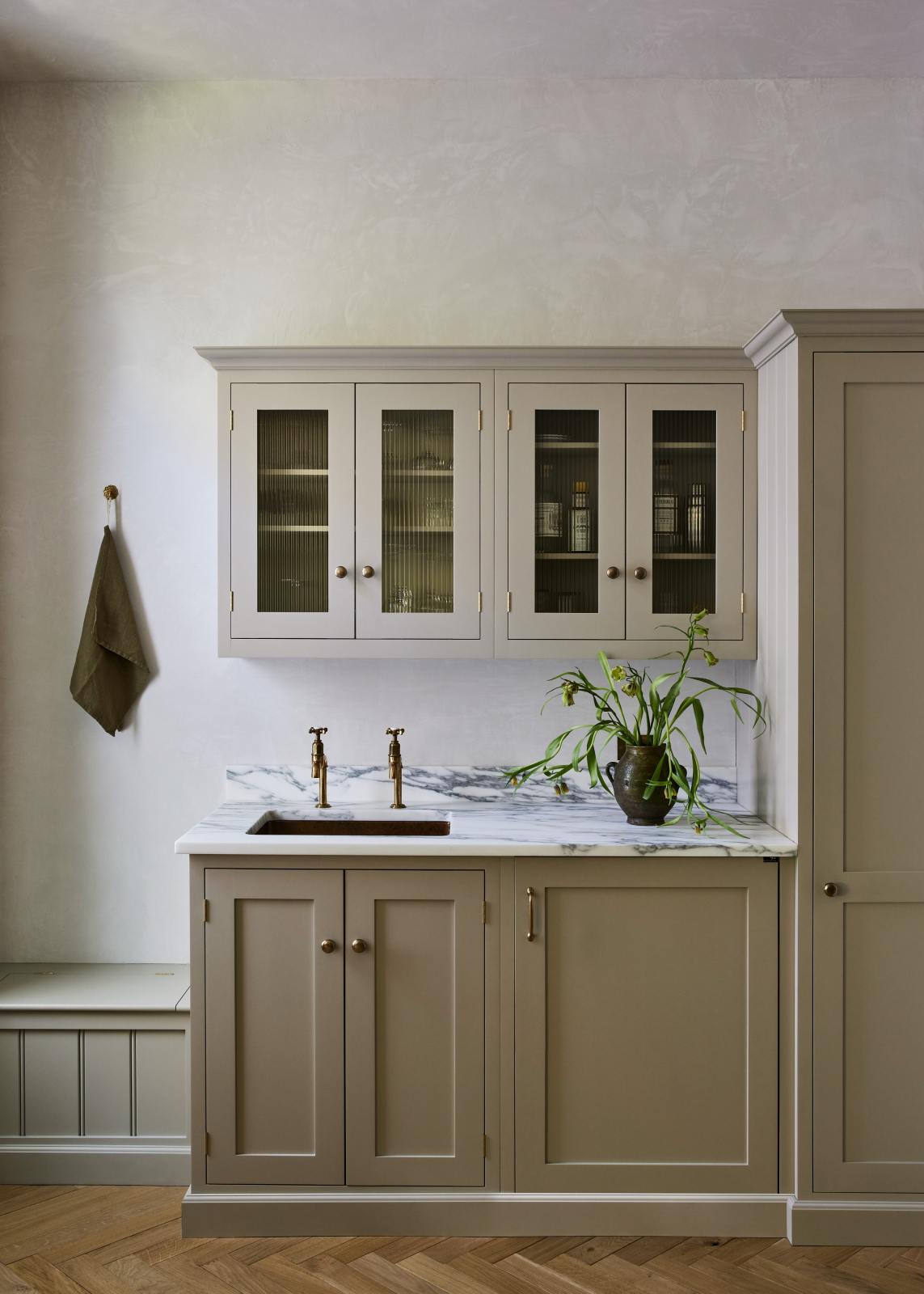 An elegant mushroom-brown, classic English kitchen features a wet bar with a single copper sink, aged brass Mayan taps, an Arabescato marble worktop, and reeded wall cupboards. Beautifully decorated with artisan ceramic and green bell flowers, it adds a sophisticated touch to the elegant kitchen.