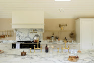 A grand mushroom-coloured classic English kitchen features a cooker run with a suspended chimney breast, a large kitchen island, two sets of sinks, aged brass Ionian taps, and an Arabescato marble worktop. Beautifully decorated with aged brass hanging rails, vintage baskets, simple ceramics, a brass folded wall lamp, and a large collection of kitchen utensils, this space creates a humble family atmosphere despite its luxurious design.