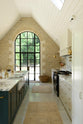 This grand mushroom-coloured classic English kitchen features a cooker run with a suspended chimney breast, a large kitchen island topped with Arabescato marble, and a bank of full-height cupboards. Beautifully decorated with woven rugs, a vintage basket, simple ceramics, brass folded wall lamps, and a large collection of kitchen utensils, this space creates a humble family atmosphere despite its luxurious design.