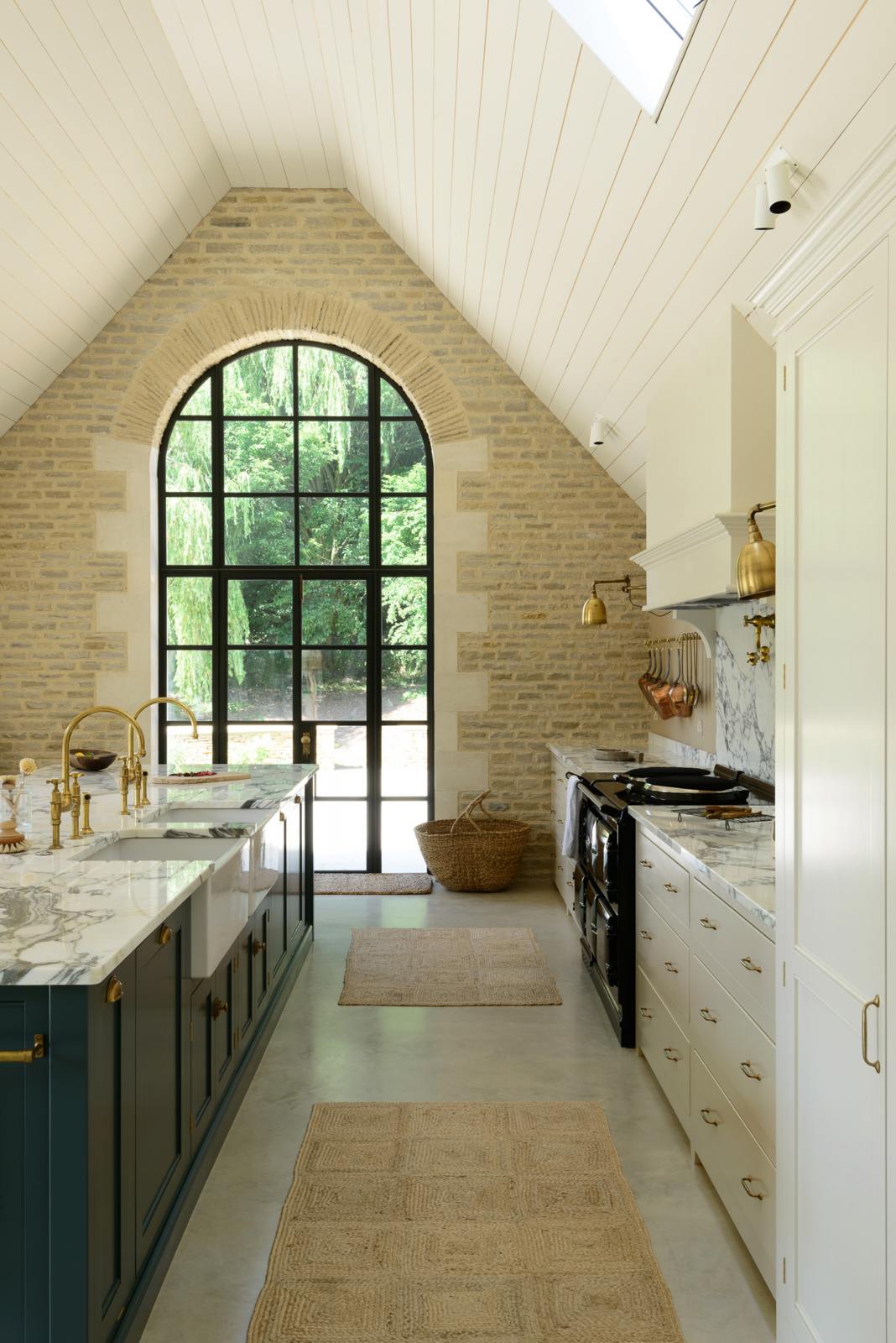 This grand mushroom-coloured classic English kitchen features a cooker run with a suspended chimney breast, a large kitchen island topped with Arabescato marble, and a bank of full-height cupboards. Beautifully decorated with woven rugs, a vintage basket, simple ceramics, brass folded wall lamps, and a large collection of kitchen utensils, this space creates a humble family atmosphere despite its luxurious design.