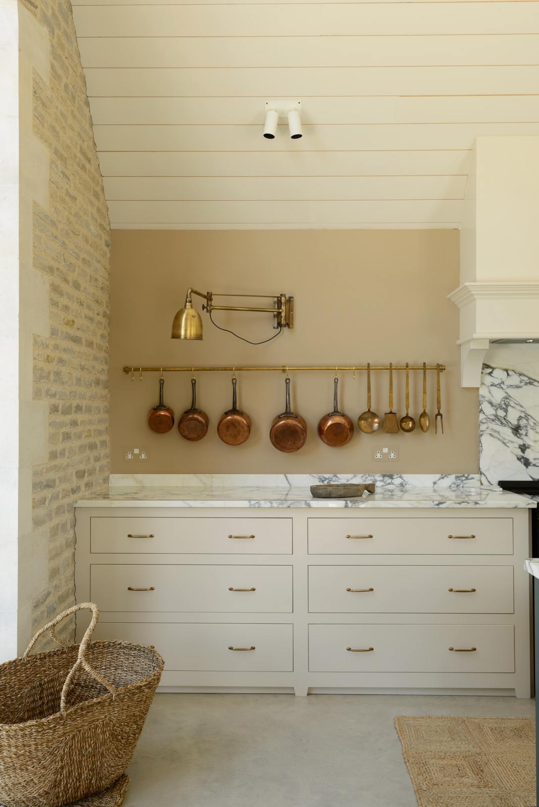 A quiet corner of a grand mushroom-coloured classic English kitchen features a cooker run with a suspended chimney breast, and an Arabescato marble worktop. Beautifully decorated with an aged brass hanging rail, a vintage basket, copper pans, a brass folded wall lamp, and a large collection of kitchen utensils, this space creates a humble family atmosphere despite its luxurious design.