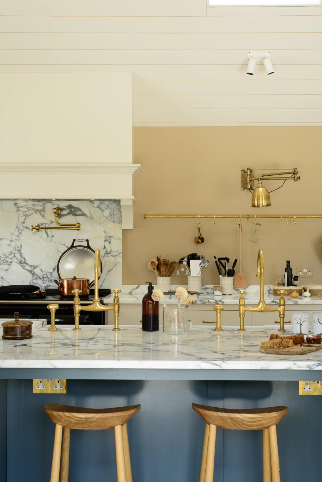 A grand mushroom-coloured classic English kitchen features a cooker run with a suspended chimney breast, a large kitchen island, two sets of sinks, aged brass Ionian taps, and an Arabescato marble worktop. Beautifully decorated with a aged brass hanging rail, bum stools, simple ceramics, a brass folded wall lamp, and a large collection of kitchen utensils, this space creates a humble family atmosphere despite its luxurious design.