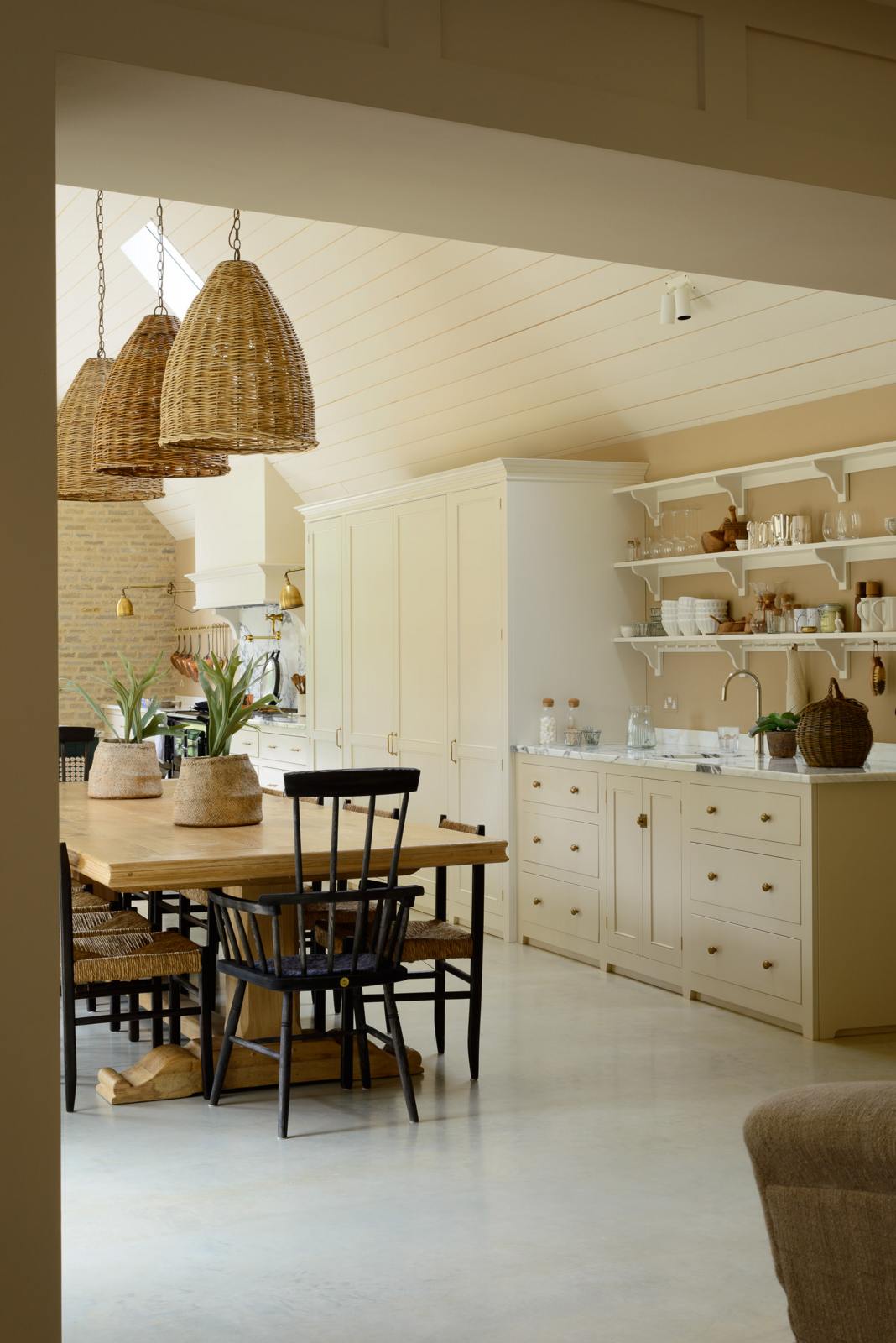 An oblique view of this grand mushroom-coloured classic English kitchen showcases a cooker run with Arabescato marble, a bank of full-height cupboards, and a small sink run with shelves. Beautifully decorated with a long dining table and woven embellishments such as pendant lights, chairs, rugs, a basket, and potted plant covers, this space creates a humble family atmosphere despite its luxurious design.