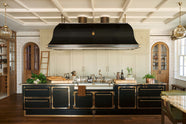 A grand room with high panelled ceiling, traditional glossy white tiled walls with curved archways on either side of a huge bank of creamy mushroom fitted full-height Classic cupboards with a polished oak ladder. An elaborate black with gold accent kitchen island and canopy sits in the middle of the space with an array of articulated brass sink taps around a wide and shallow sink.