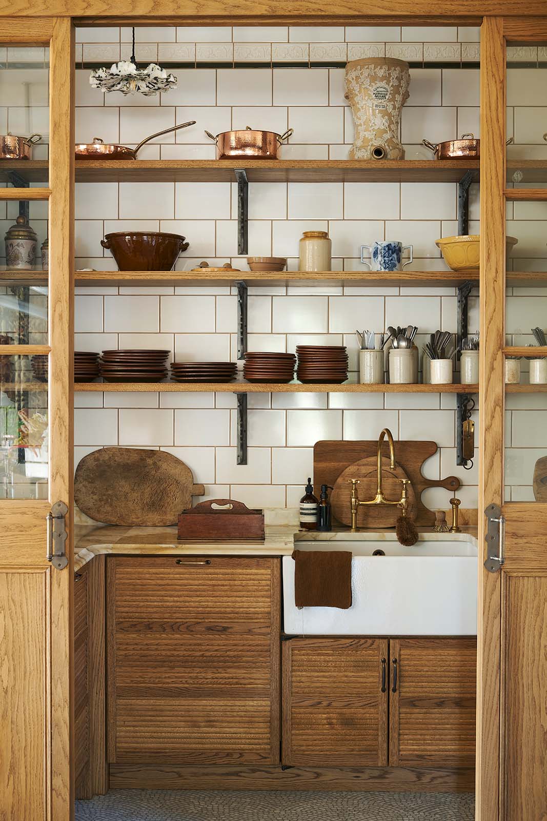 A large haberdasher’s pantry complete with its own haberdasher’s sink run, topped with a marble worktop, large shelves, and an assorted collection of ceramic plates, bowls, pots, pans, and silverware.