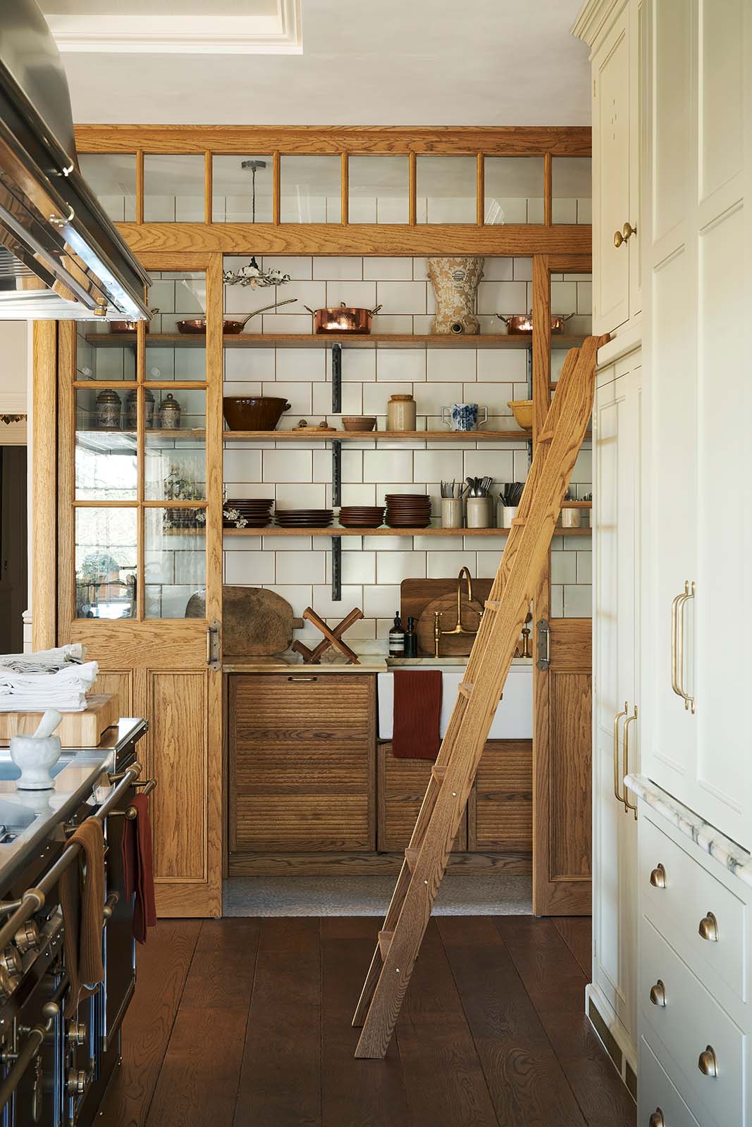 Looking down the grand kitchen with a high panelled ceiling, traditional glossy white tiled walls, and a huge bank of creamy mushroom full-height classic cupboards with a polished oak ladder is a large haberdasher’s pantry. It’s complete with its own haberdasher’s sink run topped with a marble worktop, large shelves, and an assorted collection of ceramic plates, bowls, pots, pans, and silverware.