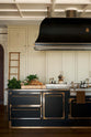 One side of a grand room with a high panelled ceiling, traditional glossy white tiled walls with curved archways on either side of a huge bank of creamy mushroom fitted full-height Classic cupboards with a polished oak ladder. An elaborate black with gold accent kitchen island and canopy sits in the middle of the space.