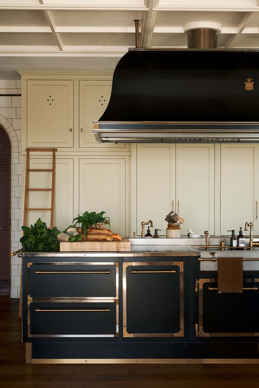 One side of a grand room with a high panelled ceiling, traditional glossy white tiled walls with curved archways on either side of a huge bank of creamy mushroom fitted full-height Classic cupboards with a polished oak ladder. An elaborate black with gold accent kitchen island and canopy sits in the middle of the space.