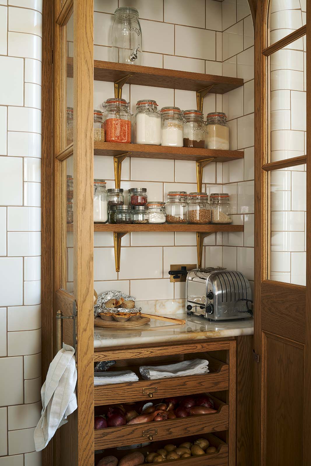 A closer, angled view reveals a small haberdasher’s pantry with tiled interior and exterior, nestled just beyond a curved archway door beside a grand wall of creamy mushroom full-height classic cupboards. The pantry features three shelves, an open base cabinet with a marble worktop, and stores colourful dried ingredients in glass jars, along with fresh vegetables and spices.