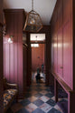A large, refectory red hallway with full-height classic cabinets, solid oak worktop, and a black Labrador lies on the red and white marble chequered floor with a toy. The ceiling displays a glass shade with copper rims and a small white porcelain wall light.