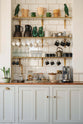 A charming personal café area features cream subway splashback tiles, three shelves with classic brass brackets, aged brass hanging rails, a Quooker Fusion mixer and instant hot tap, and a silver milk foamer. An array of simple ceramic mugs, drinking glasses, teapots, jugs, and a tray of assorted teas, coffee, and sugars completes the setup.