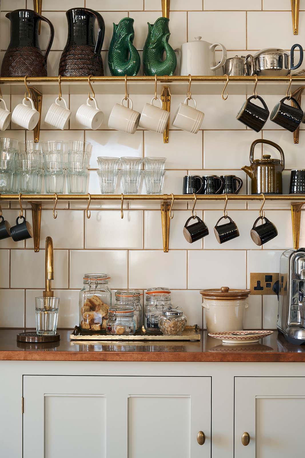 A charming personal café area features cream subway splashback tiles, three shelves with classic brass brackets, aged brass hanging rails, a Quooker Fusion mixer and instant hot tap, and a silver milk foamer. An array of simple ceramic mugs, drinking glasses, teapots, jugs, and a tray of assorted teas, coffee, and sugars completes the setup.