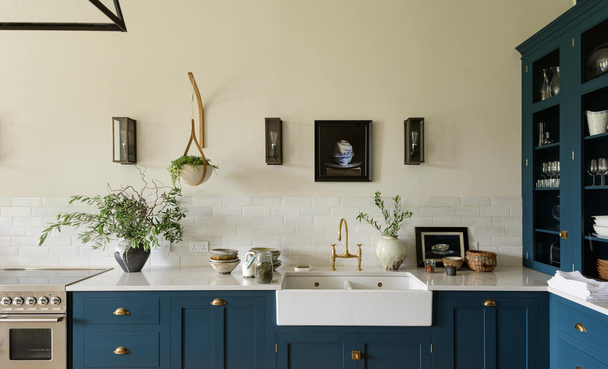 A classic English, Bond Street blue kitchen features a cooker and sink run with a Silestone worktop, glazed counter cabinet, double sink, an aged brass Ionian tap, and white crackle metro splashback tiles. The renovated medieval house is elegantly adorned with modern still-life paintings, artisan ceramics, and vibrant green plants, seamlessly blending the historical with the contemporary.
