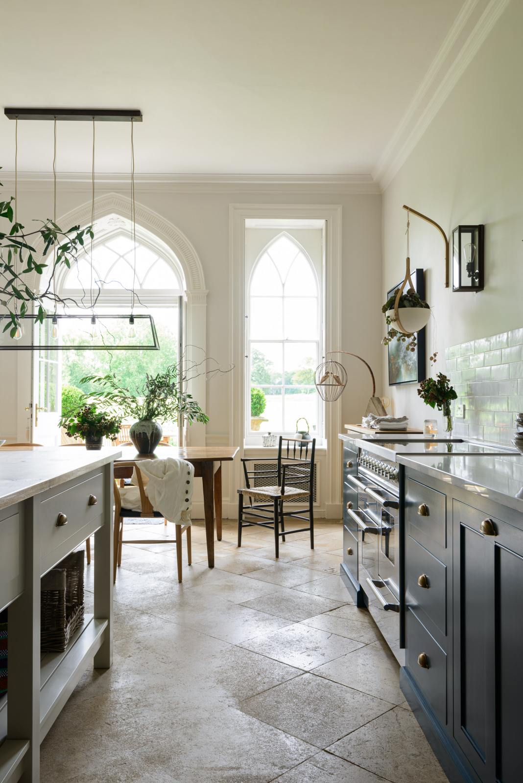 Nestled between the white linen prep table with a rustic oak worktop and the cooker and sink run with a Silestone worktop of the classic English, Bond Street blue kitchen is a minimalistic dining room with tall windows and French doors, letting in bright natural light. The renovated medieval house is elegantly adorned with modern sculptures, artisan ceramics, and vibrant green plants, seamlessly blending the historical with the contemporary.