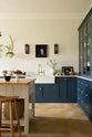 A classic English, Bond Street blue kitchen features a white linen prep table with a rustic oak worktop, a cooker and sink run with a Silestone worktop, glazed counter cabinet, double sink, an aged brass Ionian tap, and white crackle metro splashback tiles. The renovated medieval house is elegantly adorned with modern still-life paintings, artisan ceramics, and vibrant green plants, seamlessly blending the historical with the contemporary.