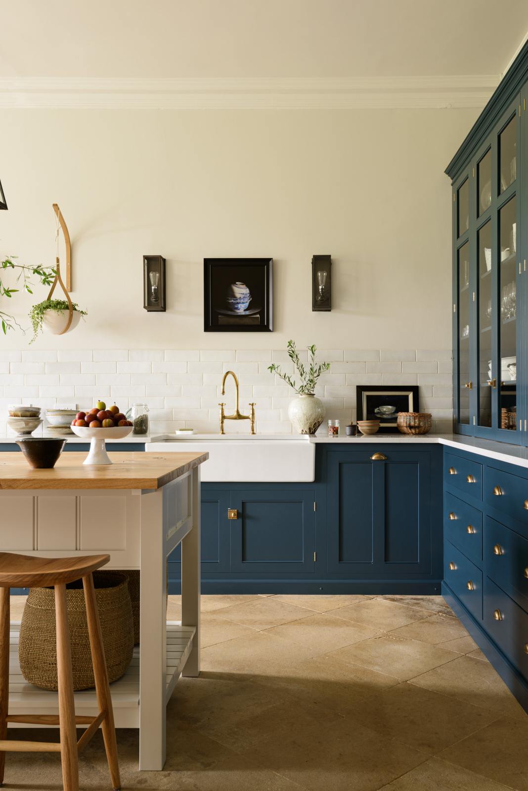 A classic English, Bond Street blue kitchen features a white linen prep table with a rustic oak worktop, a cooker and sink run with a Silestone worktop, glazed counter cabinet, double sink, an aged brass Ionian tap, and white crackle metro splashback tiles. The renovated medieval house is elegantly adorned with modern still-life paintings, artisan ceramics, and vibrant green plants, seamlessly blending the historical with the contemporary.