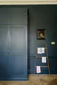 A quiet corner of a classic English Bond Street blue kitchen features a full-height cabinet and top cabinets. The renovated medieval house is elegantly adorned with modern still-life painting, a wooden rack, tea towels, and its original flooring, seamlessly blending the historical with the contemporary.
