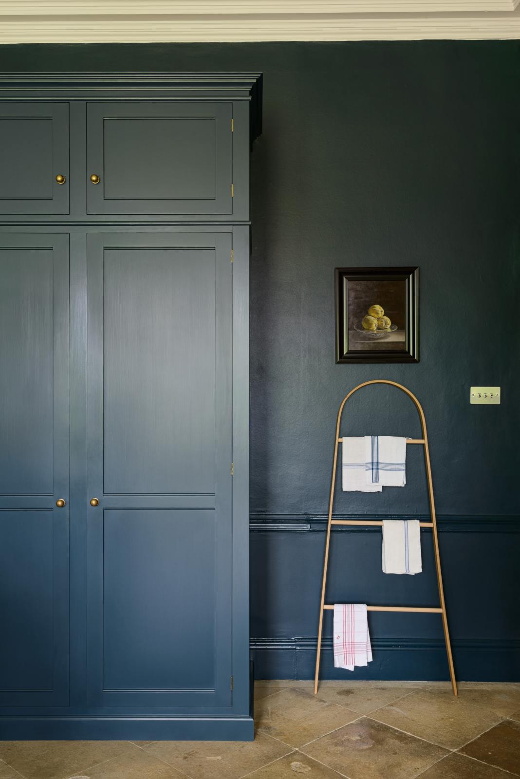 A quiet corner of a classic English Bond Street blue kitchen features a full-height cabinet and top cabinets. The renovated medieval house is elegantly adorned with modern still-life painting, a wooden rack, tea towels, and its original flooring, seamlessly blending the historical with the contemporary.
