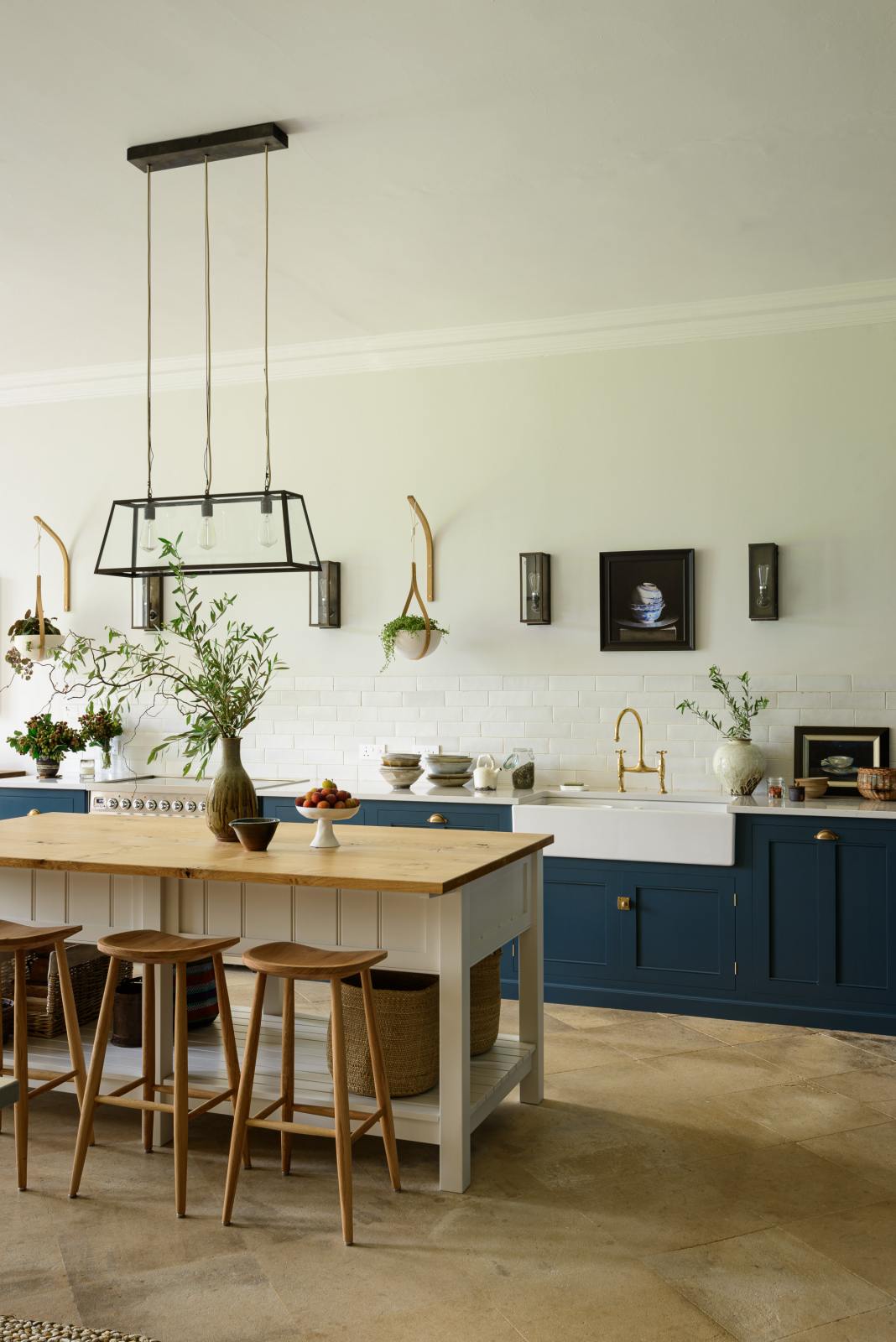 An oblique view of a classic English, Bond Street blue kitchen features a white linen prep table with a rustic oak worktop, a cooker and sink run with a Silestone worktop, a double sink, an aged brass Ionian tap, and white crackle metro splashback tiles. The renovated medieval house is elegantly adorned with modern still-life paintings, artisan ceramics, and vibrant green plants, seamlessly blending the historical with the contemporary.