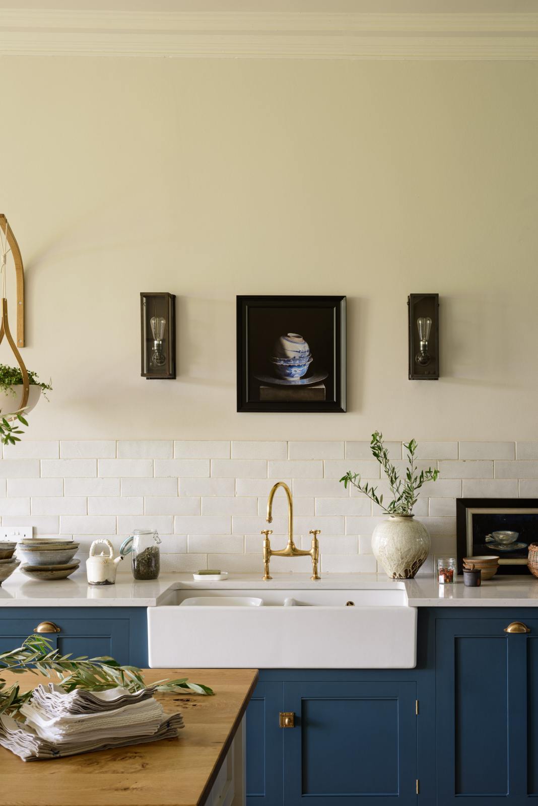 A classic English, Bond Street blue kitchen features a white linen prep table with a rustic oak worktop, a cooker and sink run with a Silestone worktop, glazed counter cabinet, double sink, an aged brass Ionian tap, and white crackle metro splashback tiles. The renovated medieval house is elegantly adorned with modern still-life paintings, artisan ceramics, and vibrant green plants, seamlessly blending the historical with the contemporary.