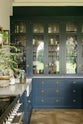 A classic English, Bond Street blue kitchen features a cooker and sink run with a Silestone worktop, glazed counter cabinets, a double sink, and an aged brass Ionian tap. The renovated medieval house is elegantly adorned with modern still-life paintings, artisan ceramics, and vibrant green plants, seamlessly blending the historical with the contemporary.