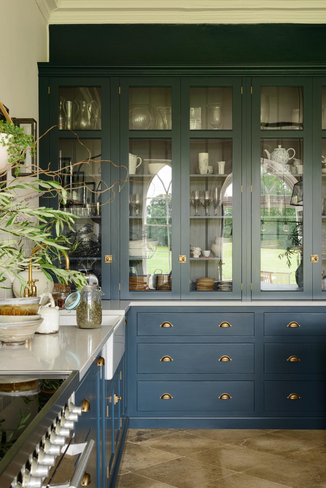 A classic English, Bond Street blue kitchen features a cooker and sink run with a Silestone worktop, glazed counter cabinets, a double sink, and an aged brass Ionian tap. The renovated medieval house is elegantly adorned with modern still-life paintings, artisan ceramics, and vibrant green plants, seamlessly blending the historical with the contemporary.