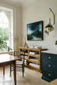 A small section of the renovated medieval house’s kitchen is a Haberdasher’s glazed drawer cabinet at the edge of the classic English Bond Street blue kitchen featuring a cooker and sink run with a Silestone worktop. It is elegantly adorned with vintage furniture, a modern painting, artisan ceramics, luscious green plants, and tablecloths, seamlessly blending the historical with the contemporary.