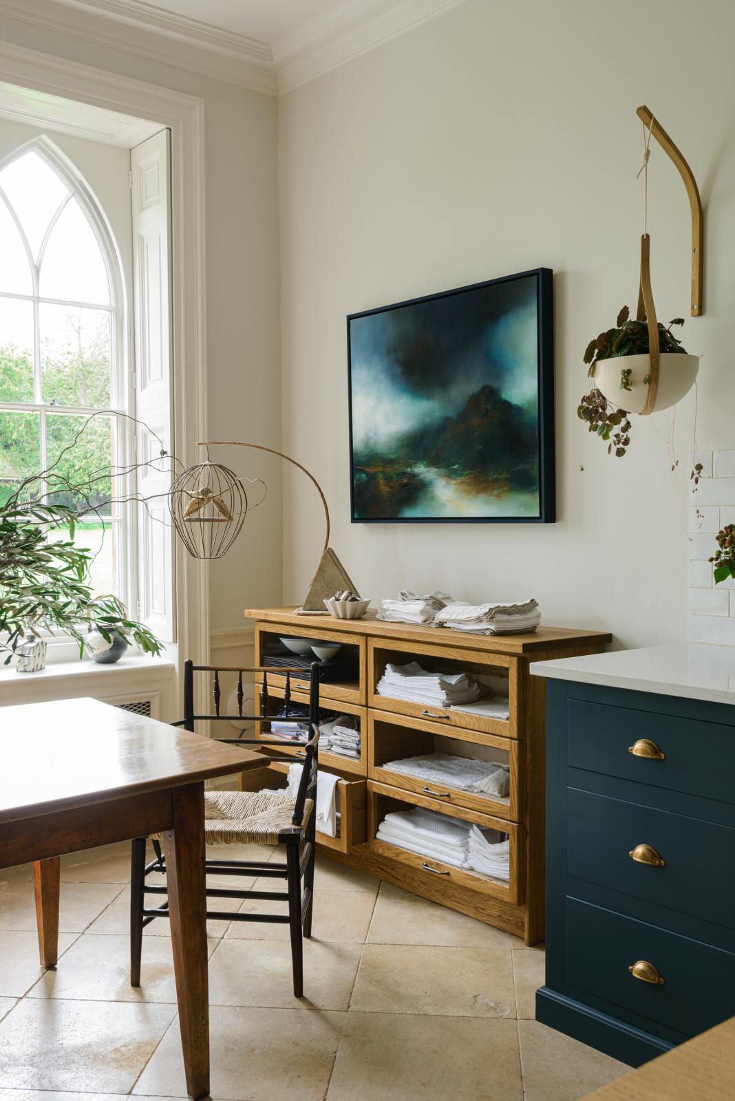 A small section of the renovated medieval house’s kitchen is a Haberdasher’s glazed drawer cabinet at the edge of the classic English Bond Street blue kitchen featuring a cooker and sink run with a Silestone worktop. It is elegantly adorned with vintage furniture, a modern painting, artisan ceramics, luscious green plants, and tablecloths, seamlessly blending the historical with the contemporary.