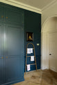 A quiet corner of a classic English Bond Street blue kitchen features a full-height cabinet and top cabinets. The renovated medieval house is elegantly adorned with modern still-life painting, a wooden rack, tea towels, and its original flooring, seamlessly blending the historical with the contemporary.