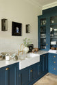 An oblique view of a classic English, Bond Street blue kitchen features a cooker and sink run with a Silestone worktop, glazed counter cabinets, a double sink, and an aged brass Ionian tap. The renovated medieval house is elegantly adorned with modern still-life paintings, artisan ceramics, and vibrant green plants, seamlessly blending the historical with the contemporary.